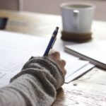 机上で勉強している手が写り、鉛筆を持っている。飲み物とノートが机上に置いてある画像